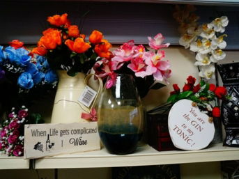 A close-up of a store shelf displaying colorful artificial flowers in vases next to decorative signs. One humorous sign reads, "When life gets complicated I Wine," and another says, "You are the GIN to my TONIC!"