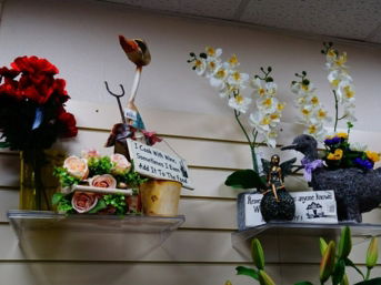 Two clear acrylic shelves mounted on a wall, showcasing novelty gifts and artificial flower arrangements. One arrangement features a quirky bird statue next to a small sign that says, "I Cook With Wine, Sometimes I Even Add It To The Food."
