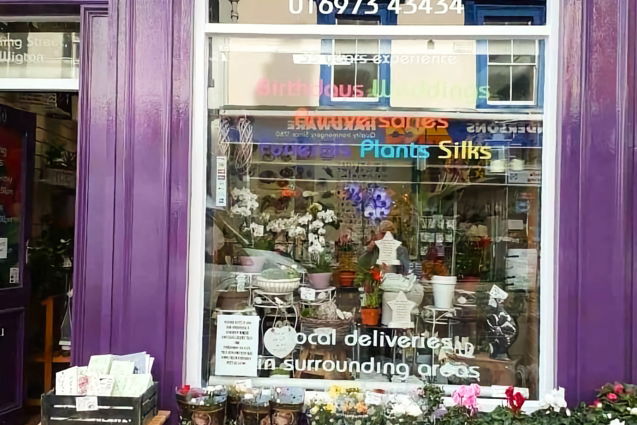 Exterior of a florist shop on King Street, Wigton. The building front is painted a bright purple. Text on the large window advertises services. The phone number is also displayed. The window is full of green plants and flowers in pots. Outside, on the sidewalk, are trays of potted flowers and a small table holding a crate of greeting cards.